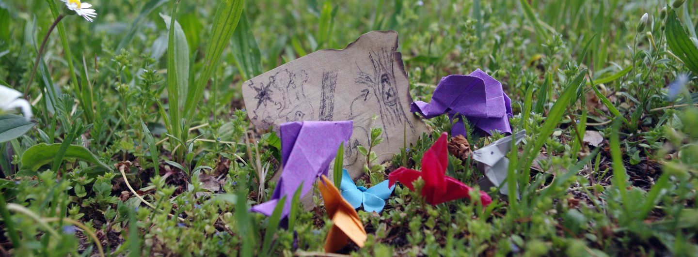 Mini-Origamis stehen im Gras und gucken auf eine kleine Schatzkarte. Ein lila Delfin, blauer Schmetterling, roter Kolibri, lila Elefant und grauer Hase.