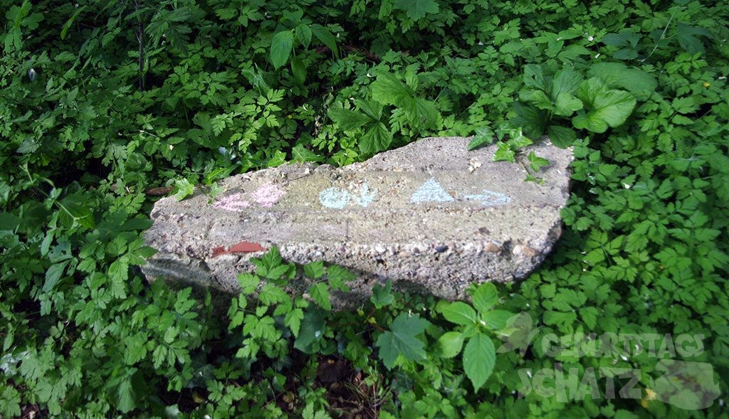 Ein großer, falcher Stein im Gebüsch mit aufgemaltem Quadrat, Kreis und Dreieck. Daneben Pfeile nach links, unten, rechts.