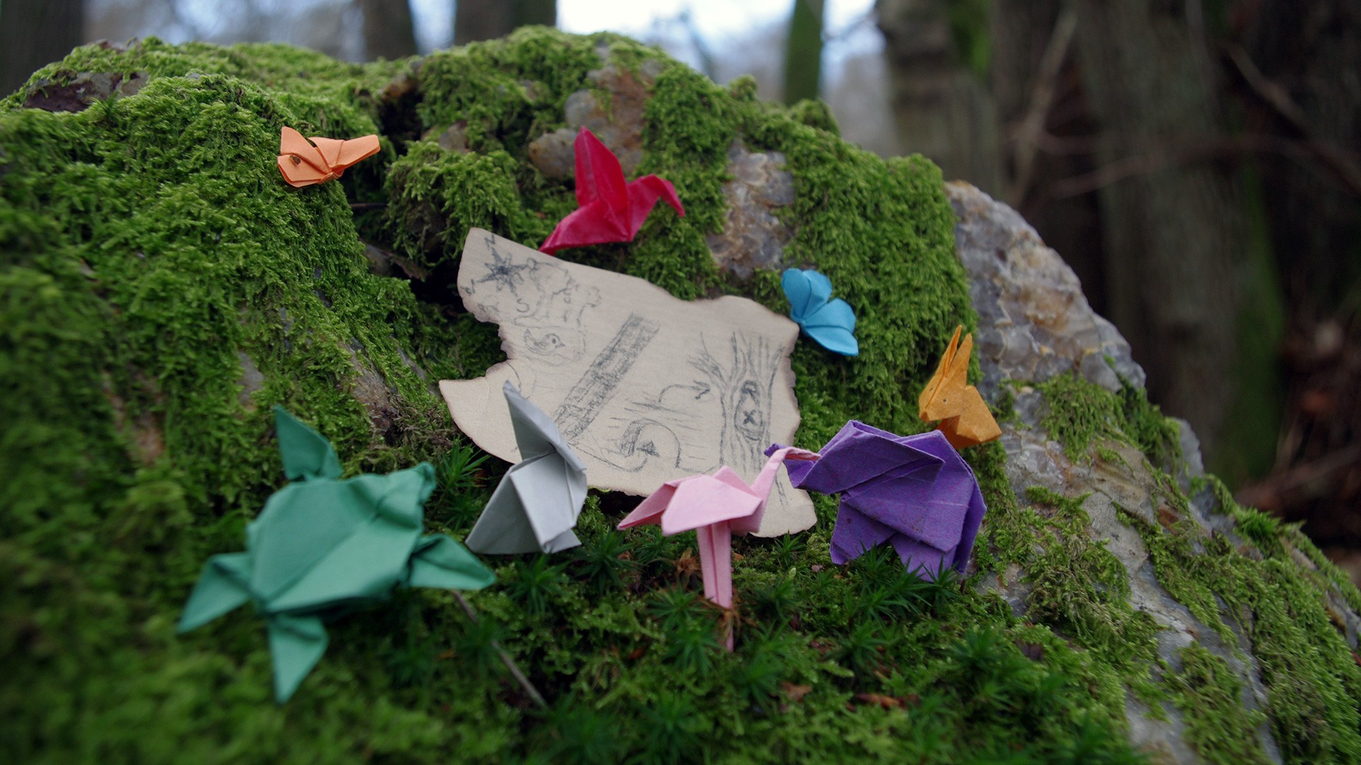 Kleine Origamis sitzen auf einem moosbedeckten Stein vor einer Miniatur-Schatzkarte.